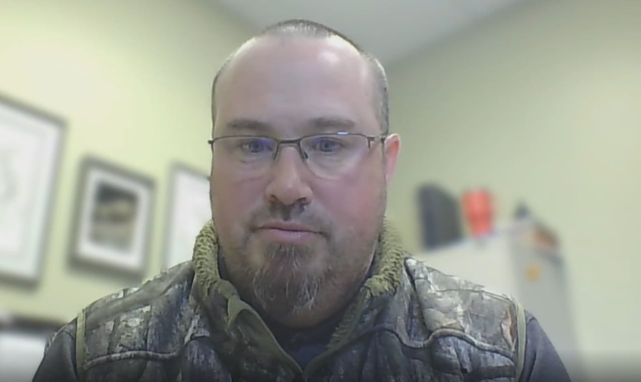 A man in a camouflage vest speaks on a video call. 