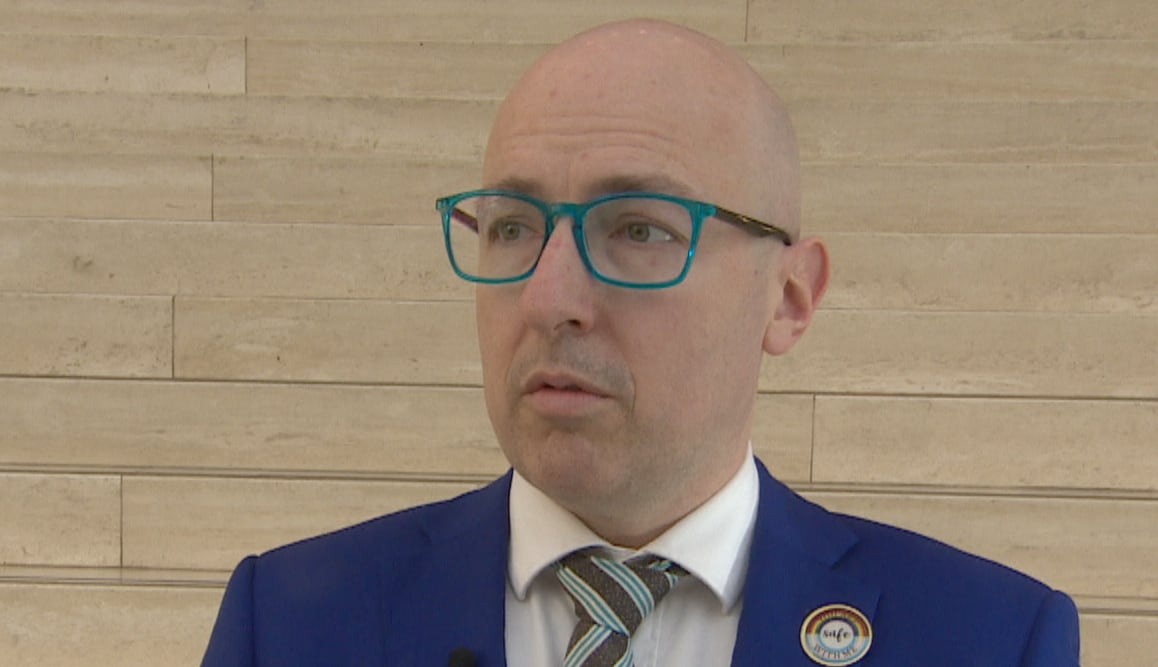A bald white man, with teal-rimmed glasses, is wearing a blue suit jacket over a white dress shirt. His time is brown-and-blue striped.