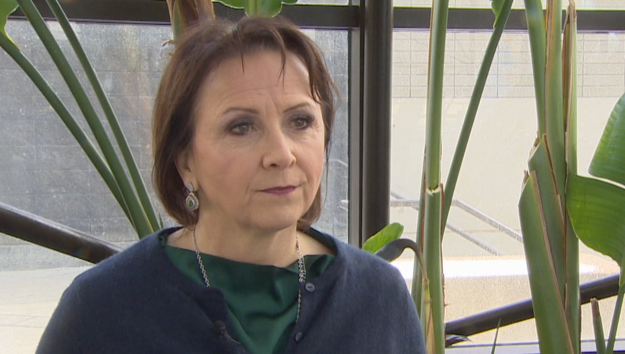 A white woman with short brown hair, wearing a navy button-up sweater over a forest-green top, is standing indoors, in front of fake greenery.