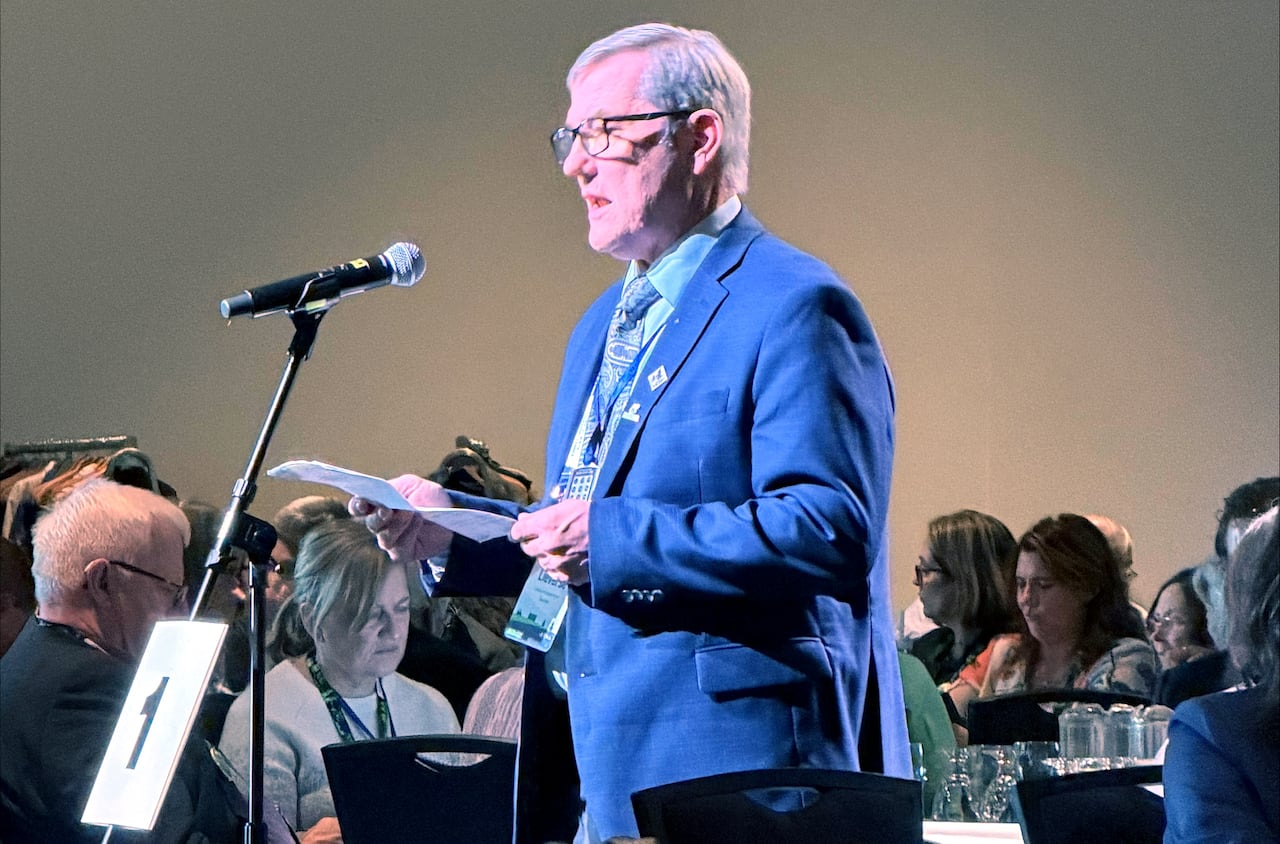 A man in a blue suit and glasses speaks into a microphone in a convention hall