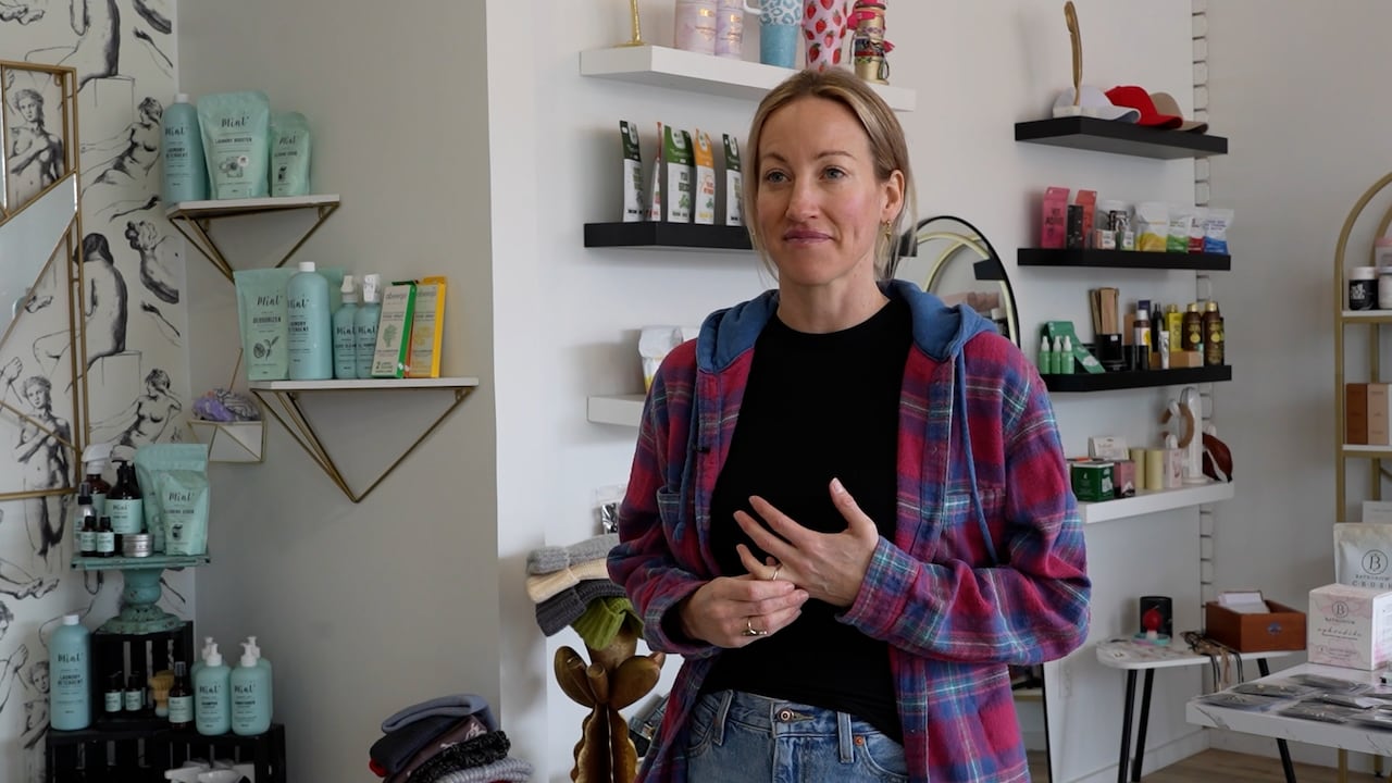 a woman in a black shirt and plaid hoodie stands in front of white walls with stocked shelves in the background. 