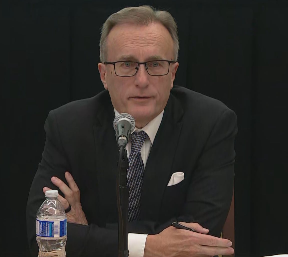A man in a suit and glasses speaks while sitting at a table.