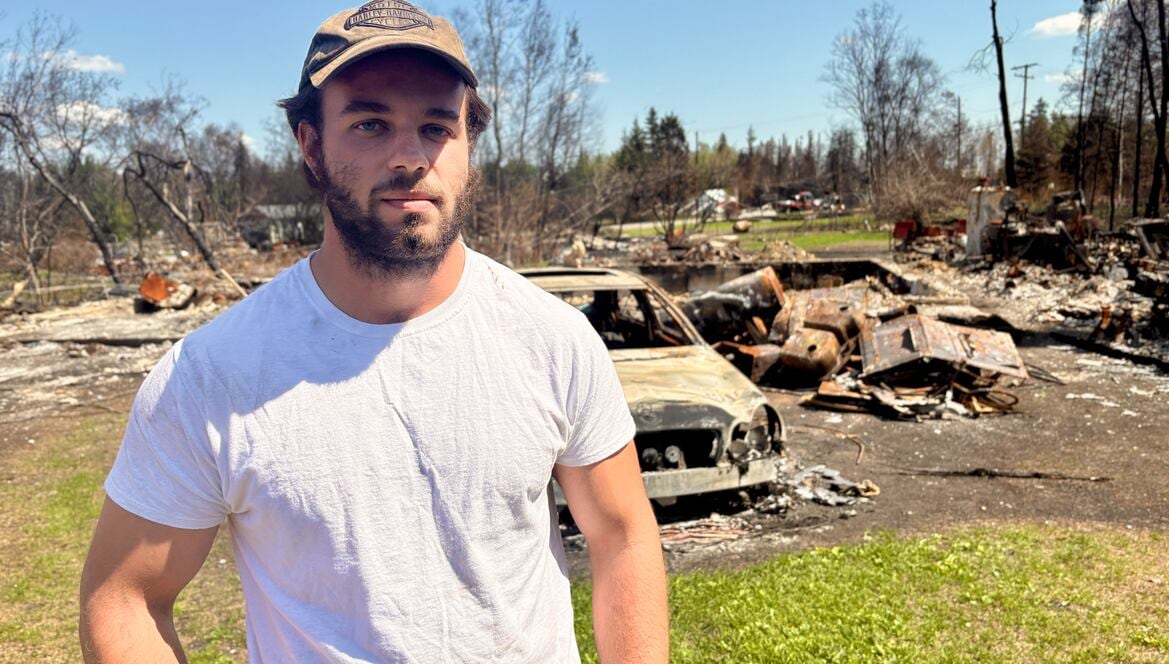 A man stands in front of a burned vehicle, homes and trees. 