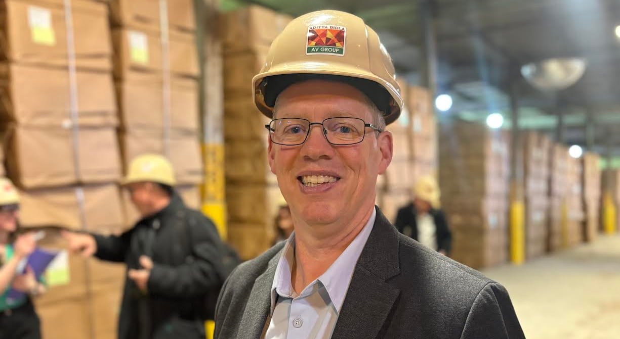 man wearing a black suit and a beige helmet smiles at the camera from inside a warehouse. 