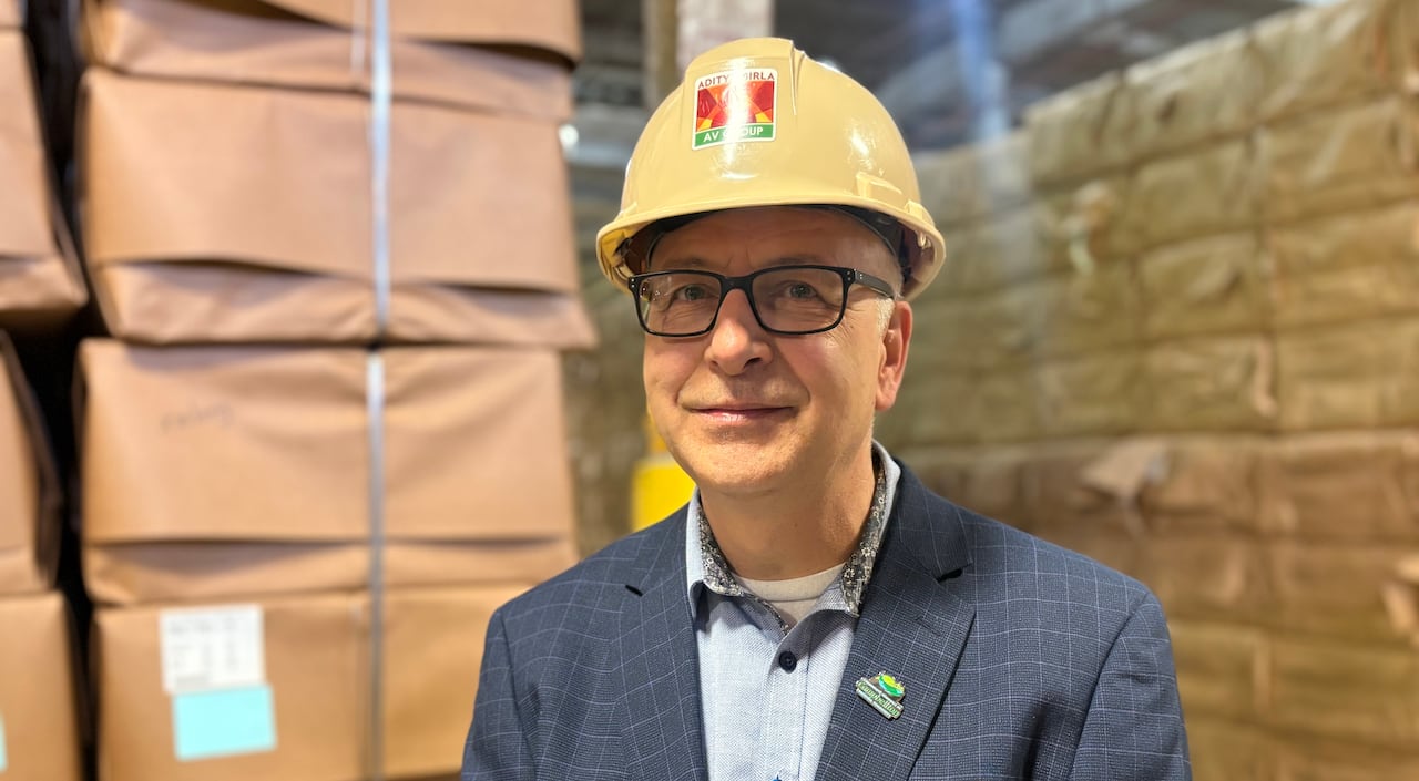 man wearing a dark blue suit and a yellow helmet stands in a warehouse smiling at the camera. 