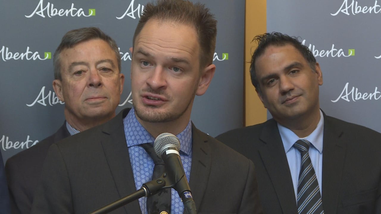 A man in a suit speaks at a microphone while two men in suits stand behind him.