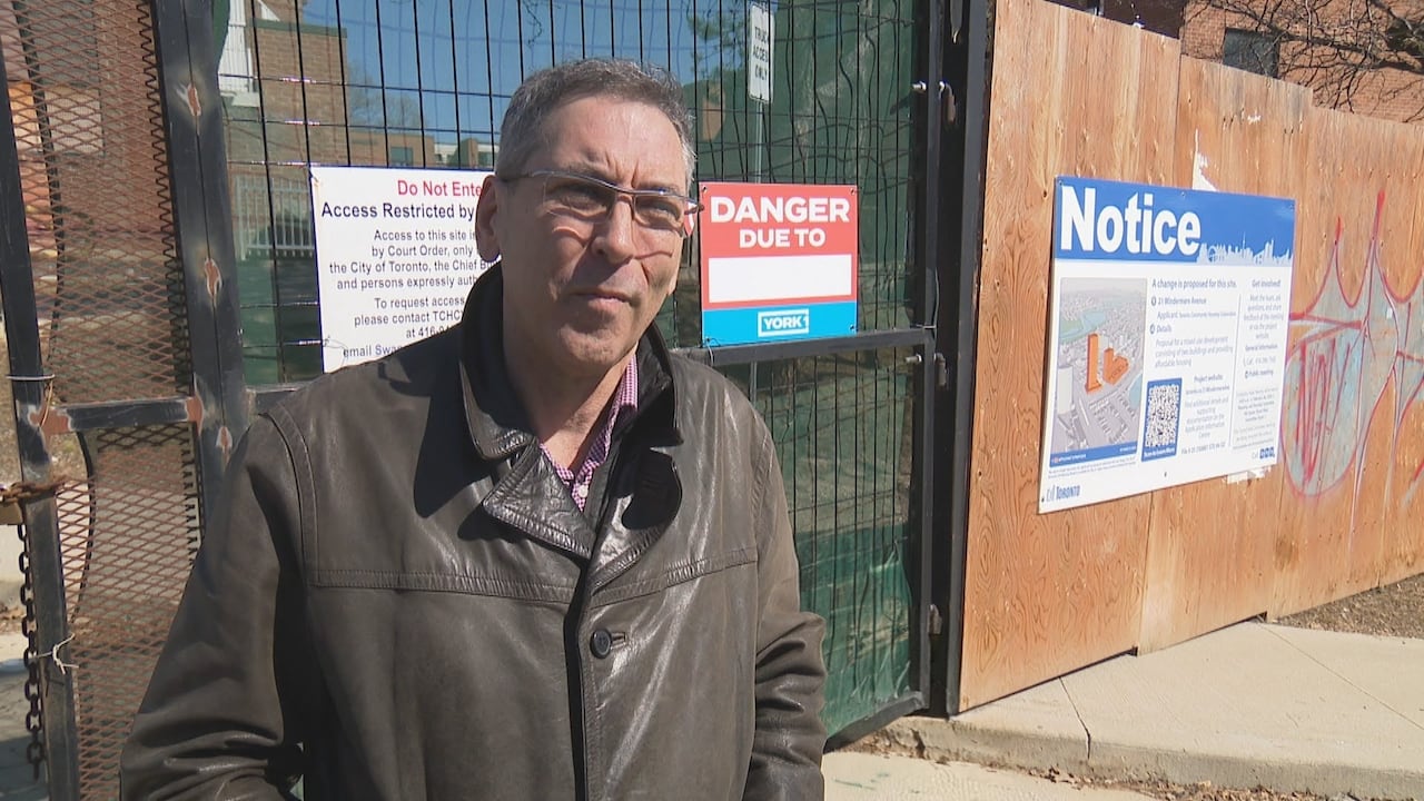 A middle-aged man wearing glasses and a leather jacket stands outside construction hoarding at the Swansea Mews site