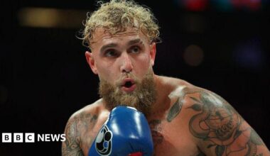 Jake Paul in action during a fight, he's shirtless and wearing blue boxing gloves.