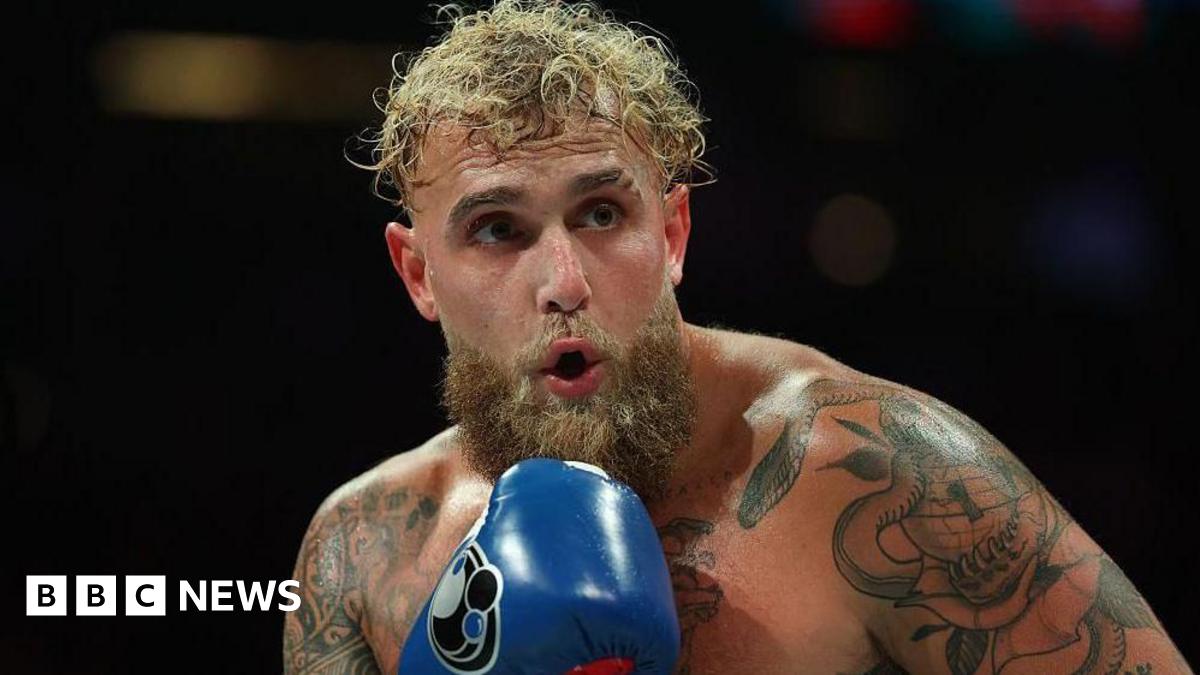Jake Paul in action during a fight, he's shirtless and wearing blue boxing gloves.