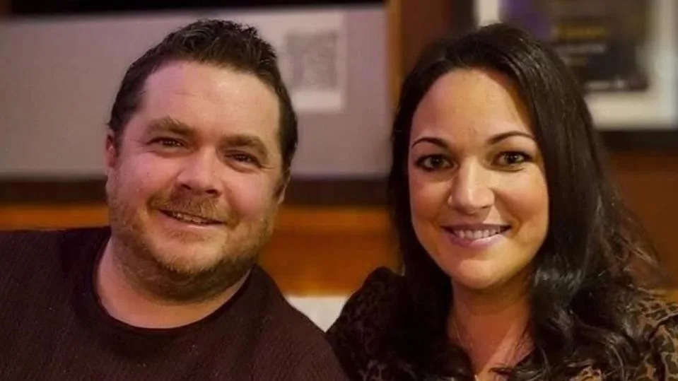 Mark and Ellen sit side  by side, smiling. Ellen has hair much darker than Mark;s and show her teeth when she smiles. She wears an animal print top. 