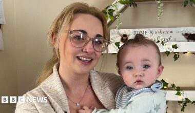 Kiely Luscombe pictured with blonde hair tied back in a pony tail. She is wearing glasses and a beige cardigan with a black top underneath. She is holding Arnold who is wearing a blue baby grow with stars on.