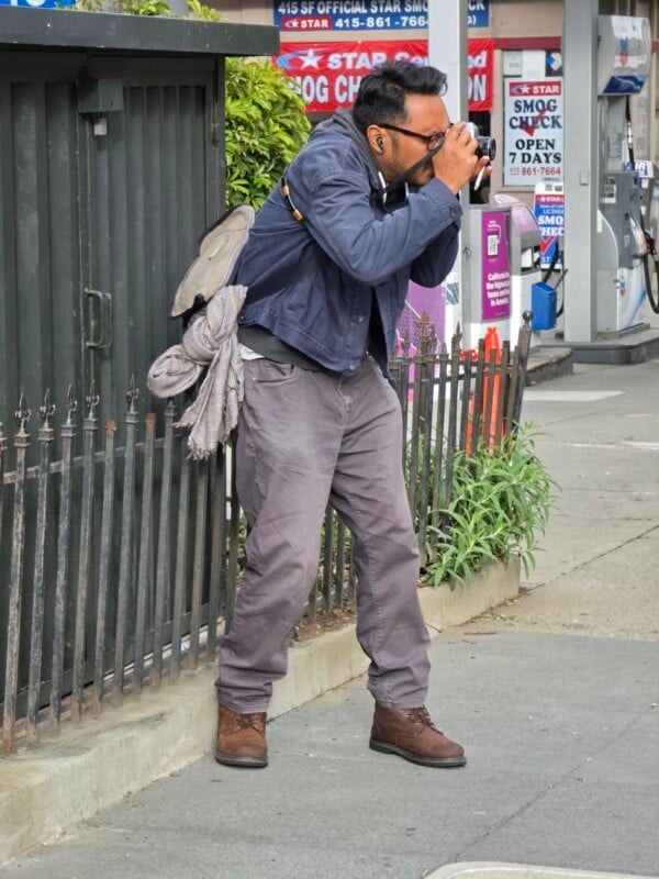 A person wearing a blue jacket and gray pants stands on a sidewalk, holding a camera up to their face, taking a photo near a metal fence with a gas station in the background.