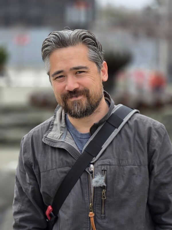 A man with salt-and-pepper hair and a beard stands outdoors, wearing a gray jacket and a shoulder strap. He is smiling softly, with a blurred urban background behind him.