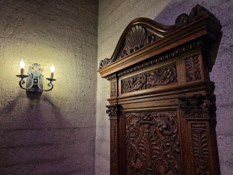 Ornate wooden panel with intricate carvings stands against a textured stone wall, illuminated softly by a nearby antique wall sconce with two exposed bulbs.