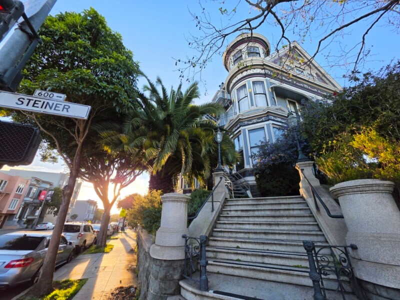 A grand Victorian-style house with a rounded turret, lush greenery, and stone steps leading to the entrance. A "Steiner" street sign is visible on the left, and cars are parked along the sunlit street.