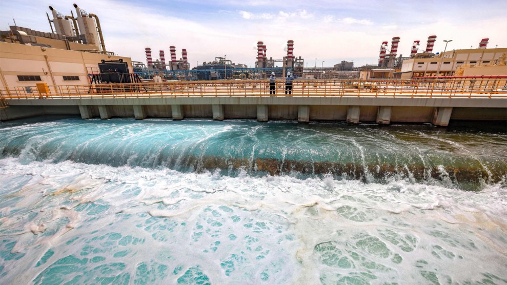 Greenish water pours through a wide sluice gate as several workers stand on a bridge across looking down at the water.