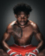 Shirtless man with a lion tattoo on his chest wears red shorts and wrapped fists. He sits intensely against a dark background.