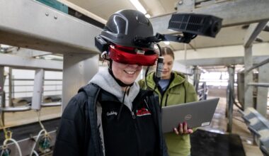 A person wearing advanced augmented reality equipment stands in a facility, while another person in the background works on a laptop.