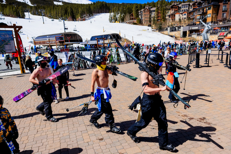 Three shirtless men wearing helmets and goggles carry ski equipment across a stone walkway at a ski resort.