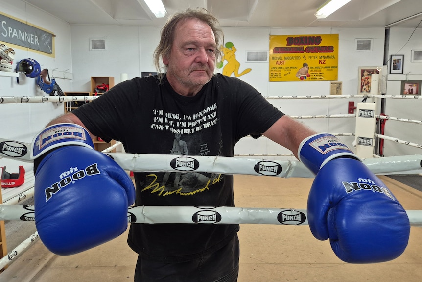 An older man with whispy hair around his face stands in a boxing ring wearing blue gloves, looking to the side of the camera.