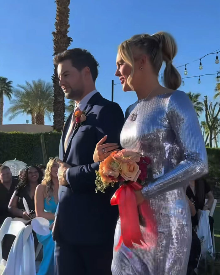 JoJo Siwa in a silver dress holding a bouquet walking down the aisle with Chris Hughes at an outdoor wedding ceremony.
