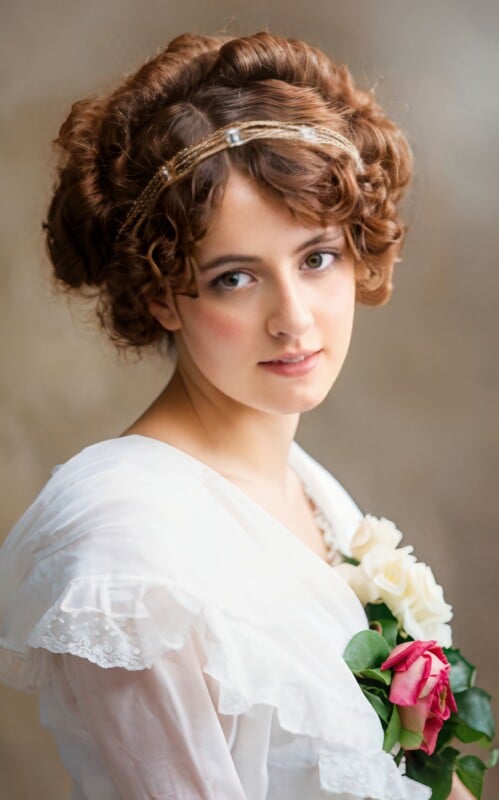 A young woman with curly brown hair, wearing a white dress and a delicate headband, holds a bouquet of white and pink roses. She looks softly at the camera against a neutral background.