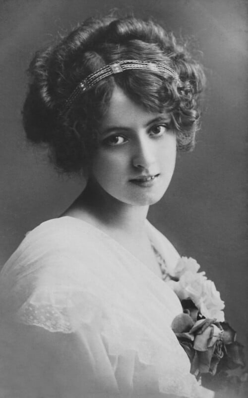 Black and white portrait of a young woman with curly hair, wearing a decorative headband and a white, lacy dress. She is gazing softly at the camera, holding a bouquet of flowers at her shoulder.