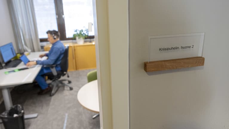 A man seen through a doorway, wearing a blue shirt and jeans sitting at a desk and looking at a laptop.