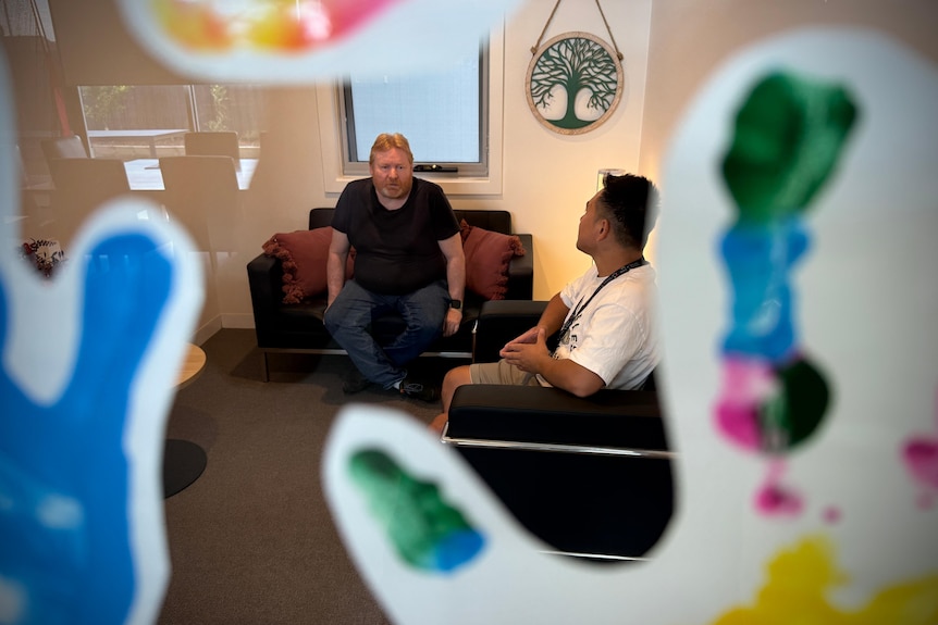 A middle-aged man, Darren Benson, and a support worker sit in chairs in a communal space.