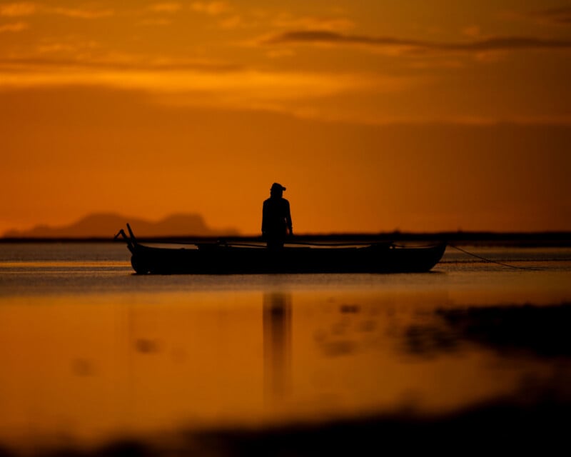 A silhouette of a person sitting in a small boat on calm water at sunset, with a vibrant orange sky and distant mountains in the background.