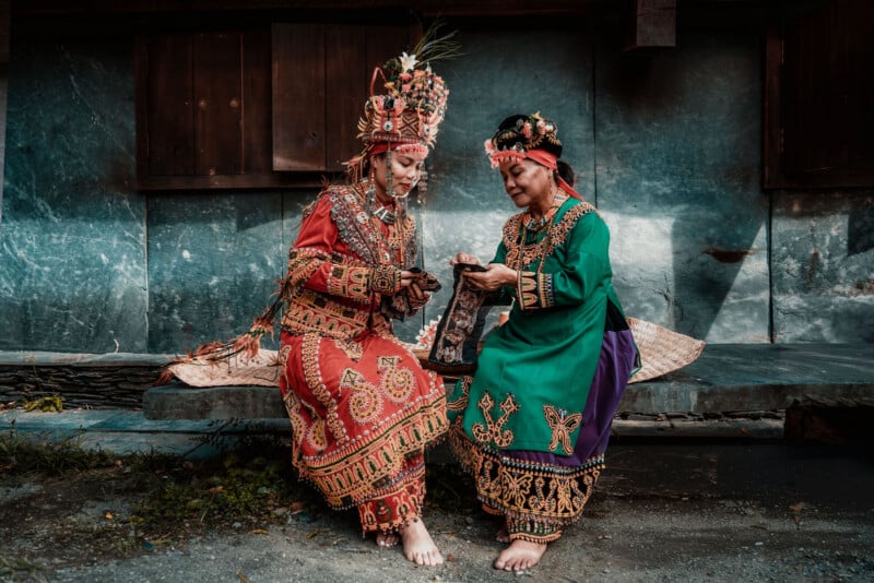 Two women in colorful, traditional attire sit on a wooden bench, focusing on an object in their hands. They wear ornate headpieces and embroidered dresses, with an old wooden building as the background.