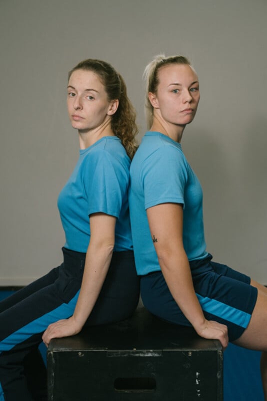 Two young women in blue athletic outfits sit back-to-back on a black box, looking directly at the camera with serious expressions against a plain background.
