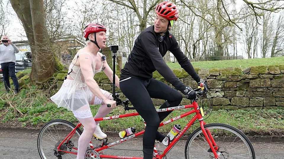 Greg in all black cycling gear on the front seat of a red tandem bike, wearing a red helmet. Jamie is on the back seat wearing white lingerie and white stockings an a red helmet.  
