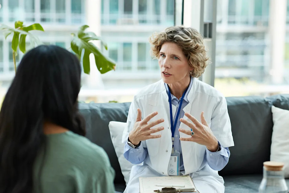 A doctor sits on a black couch speaking to a woman patient at a hospital.