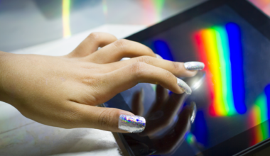 a person with light skin and sparkly long nails uses a touchscreen with a rainbow reflected on it