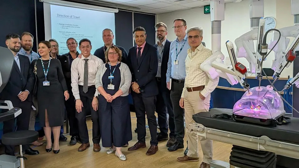 The machine looks like a plastic container with purple light inside, with lots of metal rods poking into it. Behind it are 11 people wearing smart clothing, many of them with blue NHS lanyards.