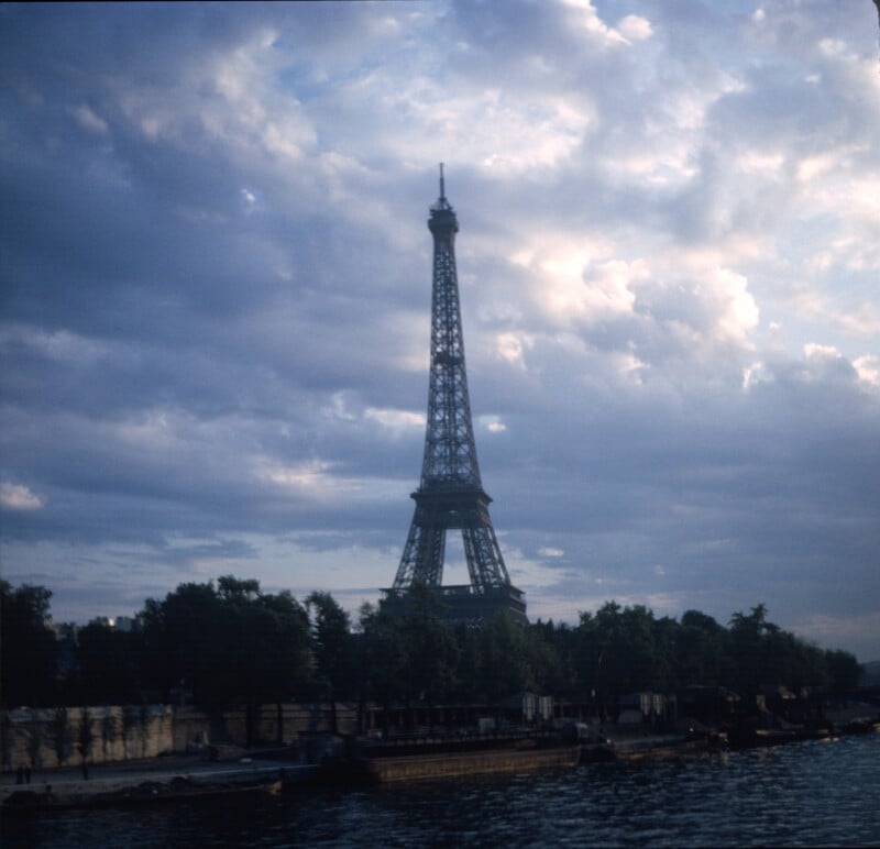 The Eiffel Tower stands against a dramatic, cloudy sky at dusk, with trees and buildings at its base and the Seine River in the foreground.