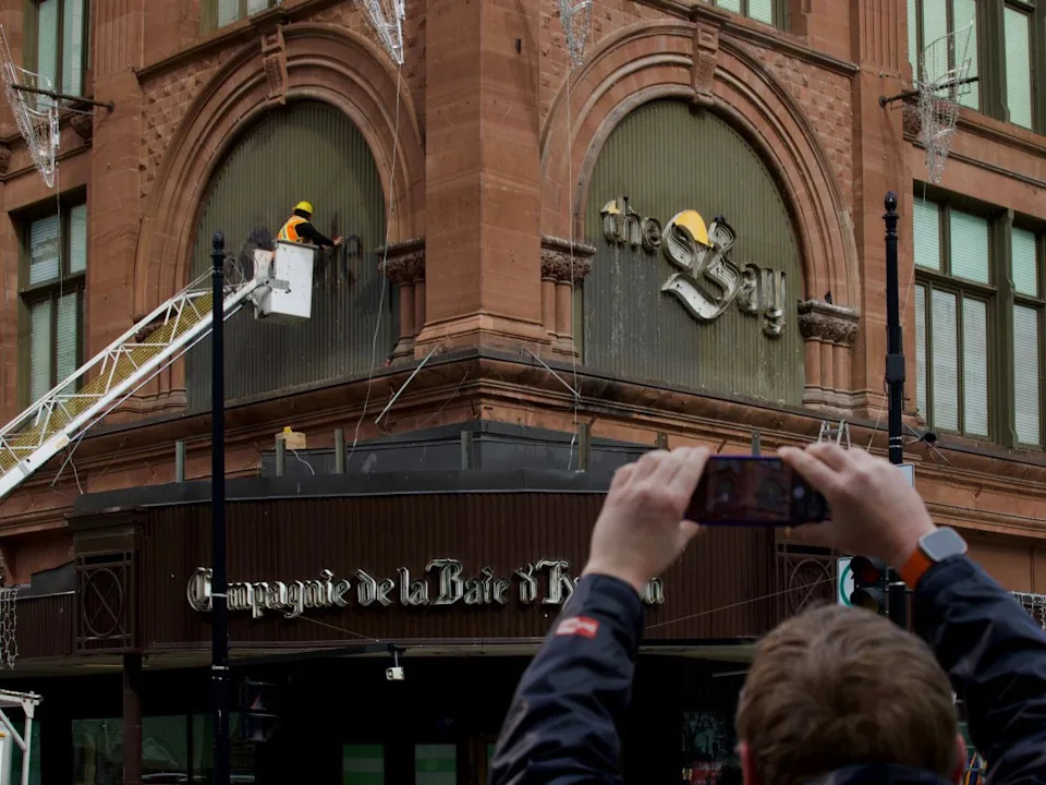  A man takes a photo of workers removing The Bay logo in downtown Montreal on Monday, March 9, 2026.