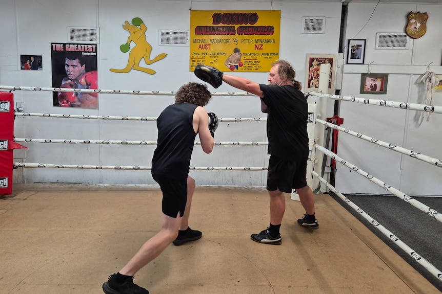 A young man with curly dark hair wearing boxing gloves and clothes ducks under the arm of an older man in the boxing ring