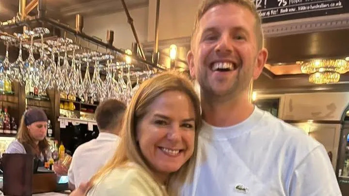 A woman and a man hug, they are in a pub, and are brother and sister.  YOu can see the bar in the background with a row of glasses and a bartender. 