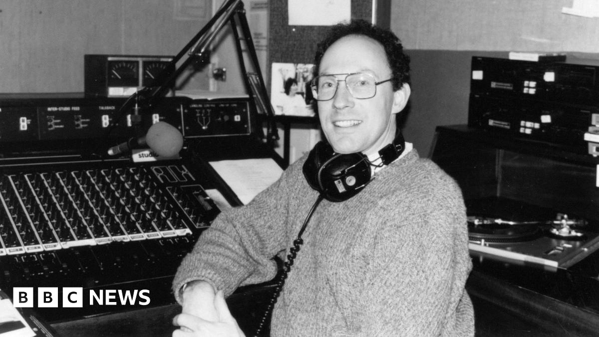 A black and white picture of a man with headphones around his neck wearing a jumper