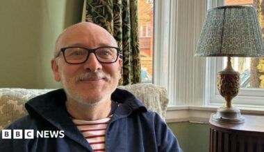 Andy Hughes sitting in front of his front window at home in Nuneaton. A lamp sits on the table behind him and neighbouring homes and parked cars can be seen outside the window in the morning sunshine.