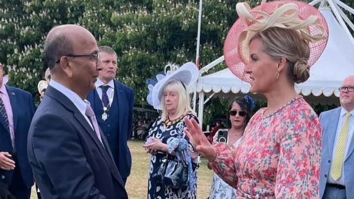A man in a dark suit speaking to a woman in a pink floral dress and hat at an outdoor formal event. Other guests in smart attire stand nearby on a grassy lawn, with a white marquee tent in the background.