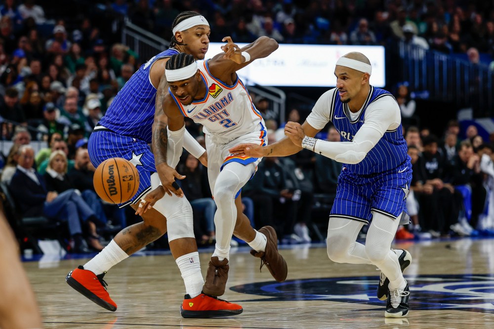 Oklahoma City Thunder guard Shai Gilgeous-Alexander (2) drives through the defense of Orlando Magic forward Paolo Banchero, left, and guard Jalen Suggs, right, during the second half of an NBA basketball game Tuesday, March 17, 2026, in Orlando, Fla. (AP Photo/Kevin Kolczynski)