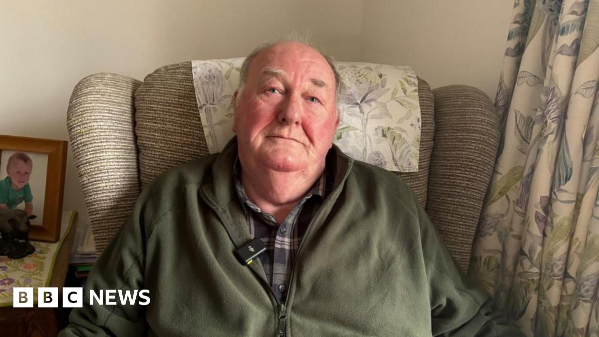 A man with grey hair sits in an arm chair wearing a checked shirt and a green zip up fleece. The arm chair is striped beige, with a patterned head rest that matches the curtain on the right of the picture. To the left, there is a picture of a young child on a side table.