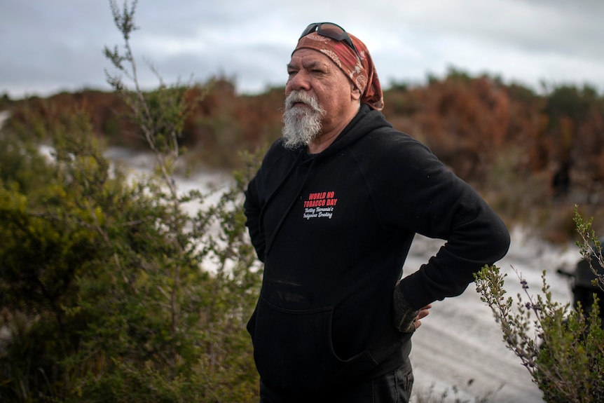 A man in a red bandana and black shirt with a grey beard stands with his hands on his hips by a sandy track in bushland. 
