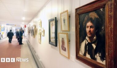 A gallery wall with various artwork in different coloured frames. The one in the foreground is a painting of a man with brown messy hair and a black suit and black tie around his white shirt. There are people further down the corridor looking at the artwork.