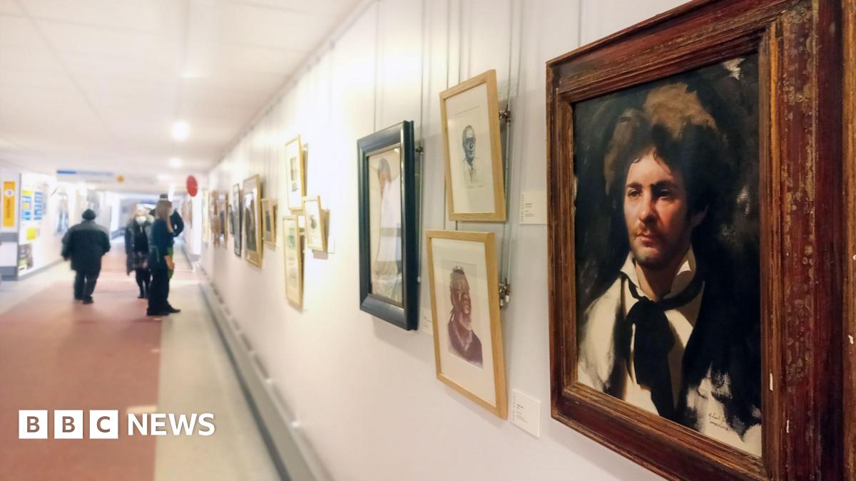 A gallery wall with various artwork in different coloured frames. The one in the foreground is a painting of a man with brown messy hair and a black suit and black tie around his white shirt. There are people further down the corridor looking at the artwork.