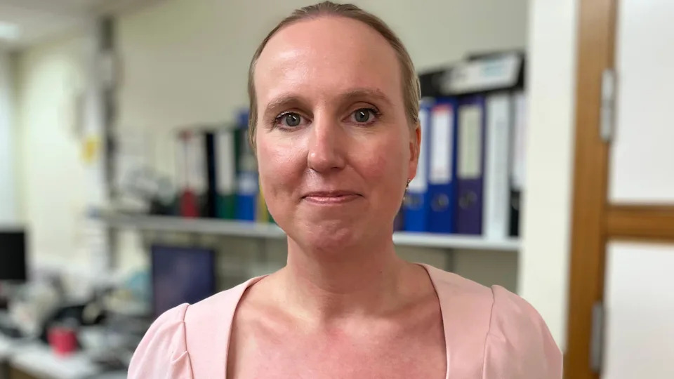 Dr Dominique Parslow smiles. She is stood in an office in a hospital. She has a pink dress on. Her blonde hair is tied back.