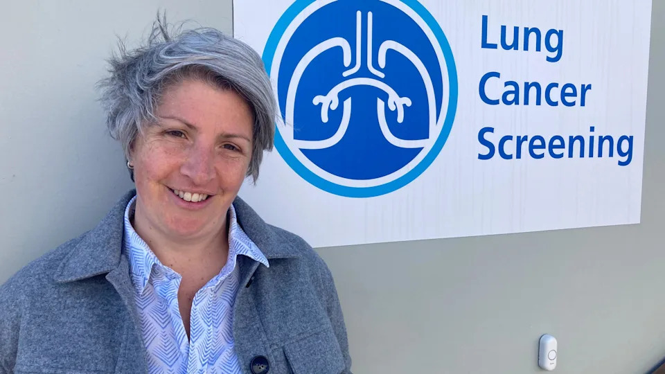 Dr Anna Bibby wearing a light shirt and grey jacket standing next to the unit which has a lung cancer screening sign on the side.